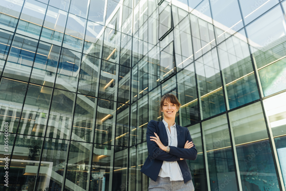 Positive female manager with crossed arms smiling while standing outside contemporary building with glass walls on city street Positive female manager with crossed arms smiling while standing outside contemporary building with glass walls on city street