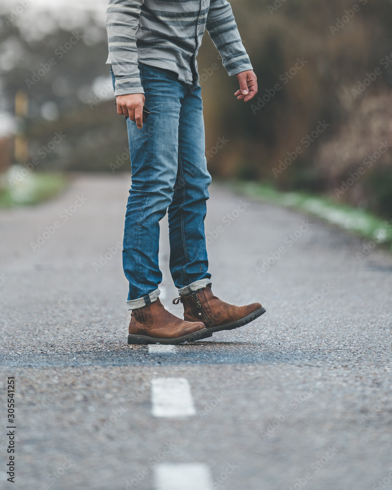 Crop unrecognizable adult male in stripped jumper and jeans walking on country roadway with hills covered by snow on background on gloomy cloudy day Crop unrecognizable adult male in stripped jumper and jeans walking on country roadway with hills covered by snow on background on gloomy cloudy day
