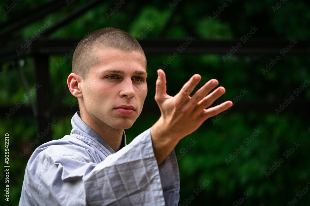Serious bald man in uniform raising arm and looking away while practicing martial arts on blurred background of gazebo and green bushes in garden Serious bald man in uniform raising arm and looking away while practicing martial arts on blurred background of gazebo and green bushes in garden