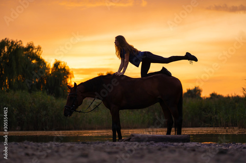 Silhouetted a slender girl practicing yoga on horseback, at sunset the horse stands in the lake. Care and walk with the horse. Strength and Beauty