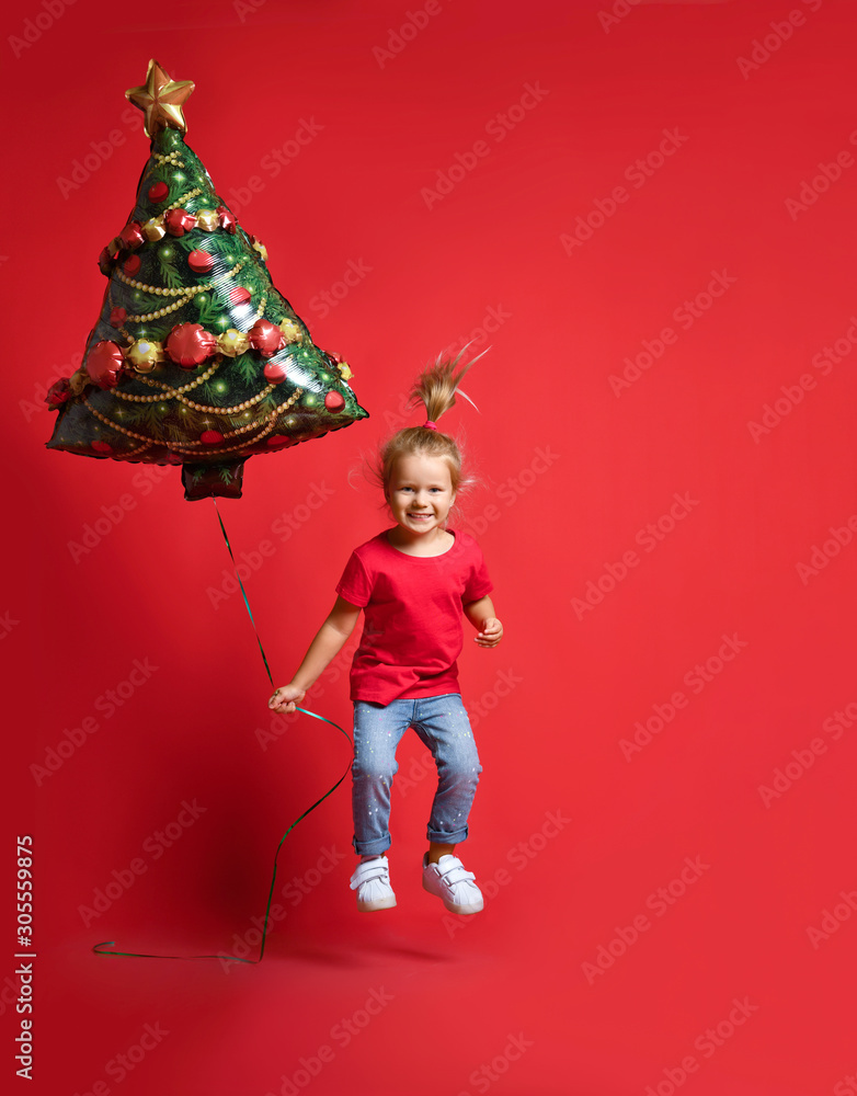 Happy toddler girl in a red T-shirt, blue jeans holding a Christmas tree balloon on a red background with free copy space.