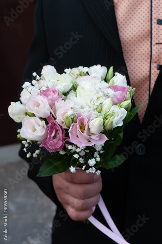 cropped view of groom in black tuxedo holding bridal bouquet with white and pink flowers with ribbon near house