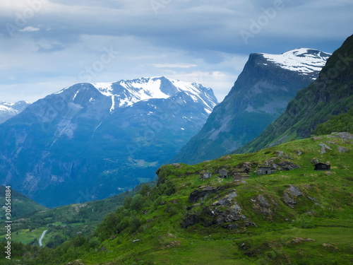 Rural wooden buildings high in the mountains of Norway blend into the green grass and bare rock surroundings in this spectacular scene, with snow on the peaks near Geiranger on a cloudy day.