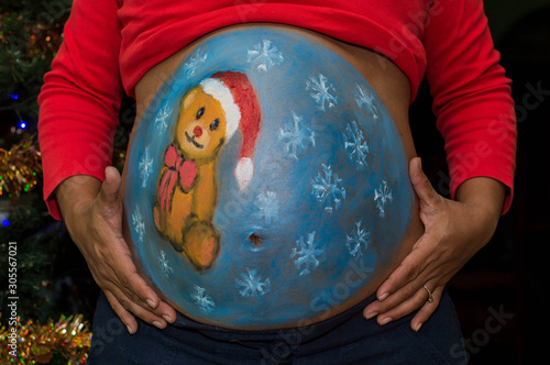 Pintura rugosa del vientre de mujer embarazada. Pintura del oso de peluche en el vientre de la mujer embarazada. Fondo con árbol de navidad photo