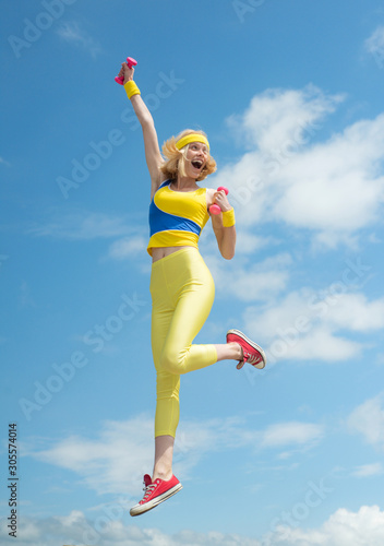 Young sporty excited girl jumping happily with hands up demonstrating exercises with pink dumbbells on sky background outdoor, panorama, copy space, sunrise light. Fashion and Trends in Sport. Health