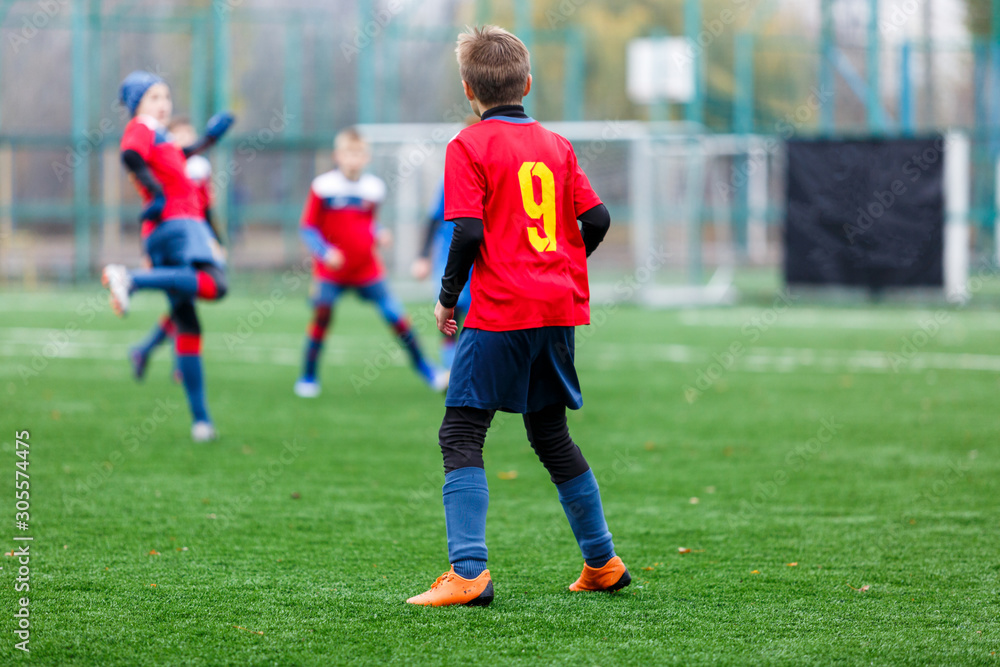 Boys in red and blue sportswear plays football on field, dribbles ball. Young soccer players with ball on green grass. Training, football, active lifestyle for kids concept 