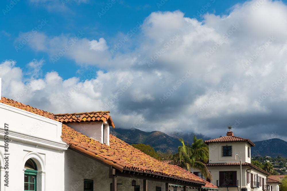 Red tile roof and white Spanish style buildings beneath palm trees, mountains, and dramatic white fluffy clouds in Santa Barbara, California