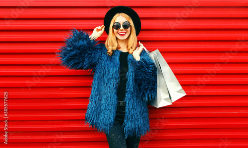 Portrait stylish smiling woman with shopping bags wearing blue faux fur coat, black round hat and sunglasses posing over red wall background © rohappy