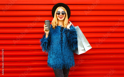 Portrait stylish smiling woman with coffee cup and shopping bags wearing blue faux fur coat, black round hat and sunglasses posing over red wall background © rohappy