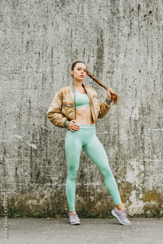 Outdoor portrait of young beautiful fit woman, wearing green activewear, athlete model posing next to grey urban wall background, sport fashion