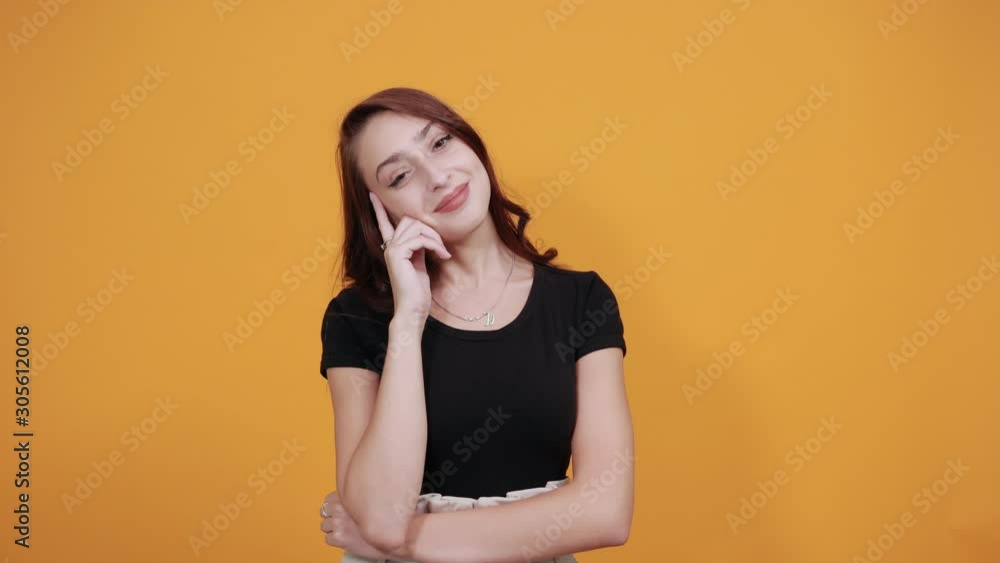 Attractive young woman in black shirt keeping finger on cheeks, looking directly isolated on orange background in studio. People sincere emotions, health concept. Attractive young woman in black shirt keeping finger on cheeks, looking directly isolated on orange background in studio. People sincere emotions, health concept.