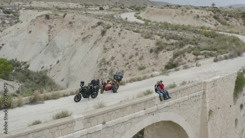 Motorcycle couple relaxing sitting on a bridge whilst touring on their bikes through Europe. Aerial panning right. photo