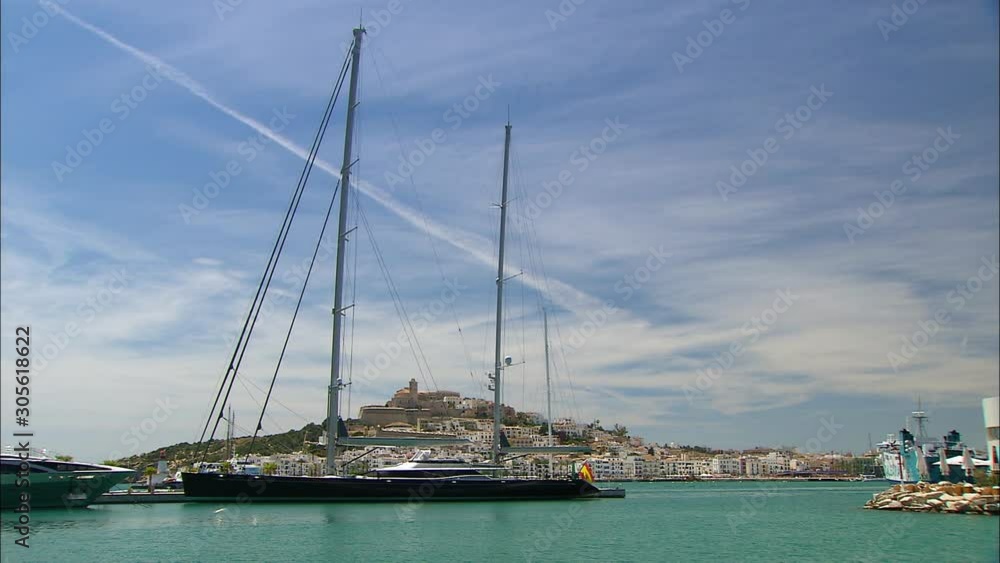 Wide low-angle still shot of a hilly Ibiza island with Spanish white apartment blocks from behind sailing and docked ships, Ibiza Balearic Island , Spain Wide low-angle still shot of a hilly Ibiza island with Spanish white apartment blocks from behind sailing and docked ships, Ibiza Balearic Island , Spain