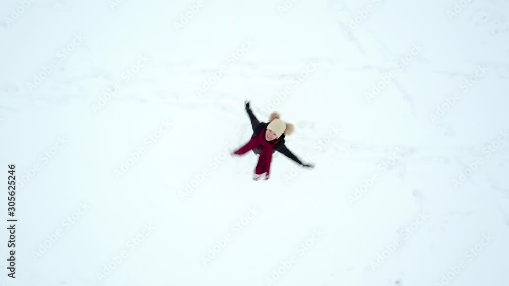 Young cheerful woman stay on snow field, look up and spin around, top-down aerial dronie shot, camera fly up while looking straight down and rotate. Girl smile and whirl, then lie and make snow angel Young cheerful woman stay on snow field, look up and spin around, top-down aerial dronie shot, camera fly up while looking straight down and rotate. Girl smile and whirl, then lie and make snow angel