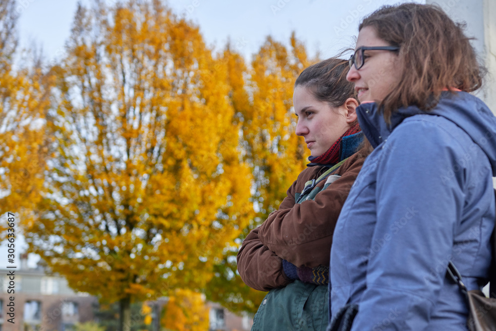 two girls looking at someone, it's autumn, there's a big yellow tree in the background