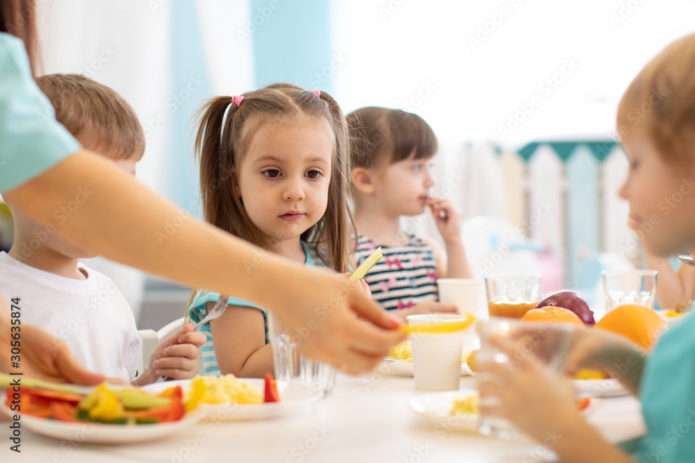 Little boys and girls from the group of preschool children sit at table with lunch and eat appetizing in kindergarten. Kids with caregiver in day care centre Little boys and girls from the group of preschool children sit at table with lunch and eat appetizing in kindergarten. Kids with caregiver in day care centre