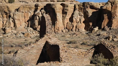 Panning across Anasazi ruins at Chetro Ketl, a religious site used thousands of years ago in Chaco Canyon, New Mexico. The brick buildings blend in with the rocky desert cliffs behind them. Shadows.