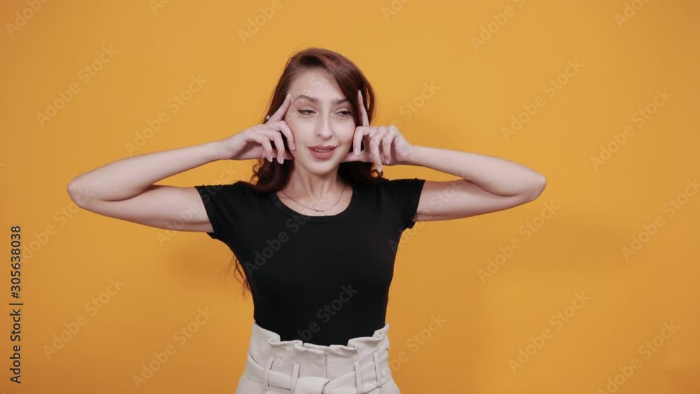 Attractive young woman in black shirt keeping finger on cheeks, doing model gesture isolated on orange background in studio. People sincere emotions, health concept. Attractive young woman in black shirt keeping finger on cheeks, doing model gesture isolated on orange background in studio. People sincere emotions, health concept.