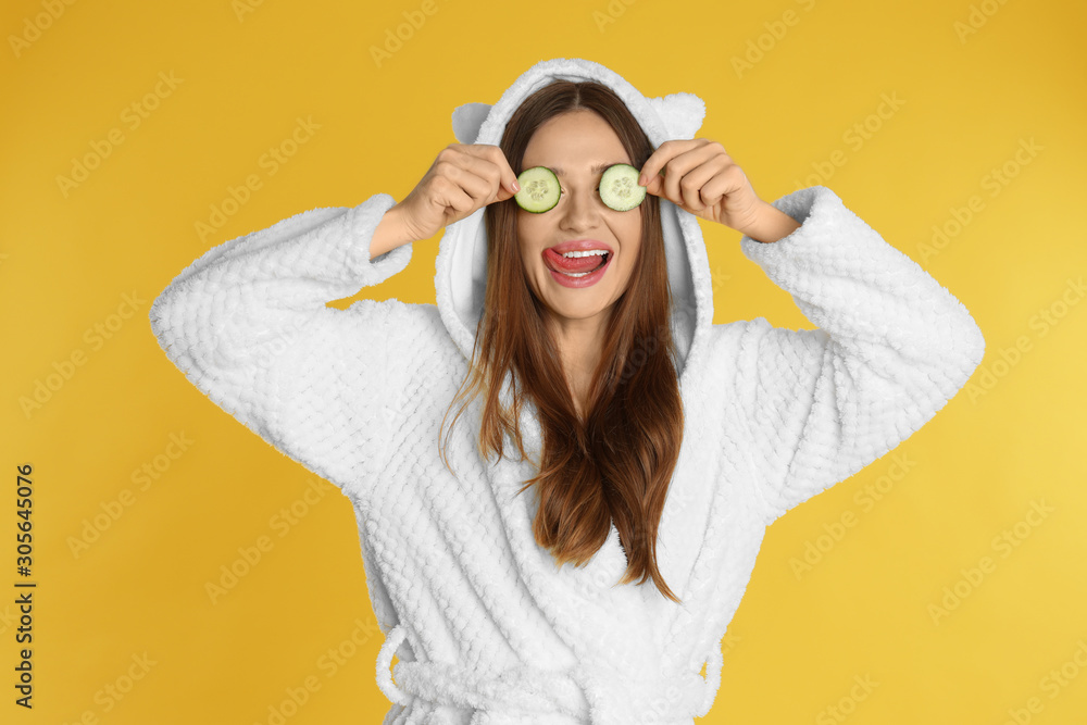 Young woman in bathrobe with cucumber slices on yellow background Young woman in bathrobe with cucumber slices on yellow background