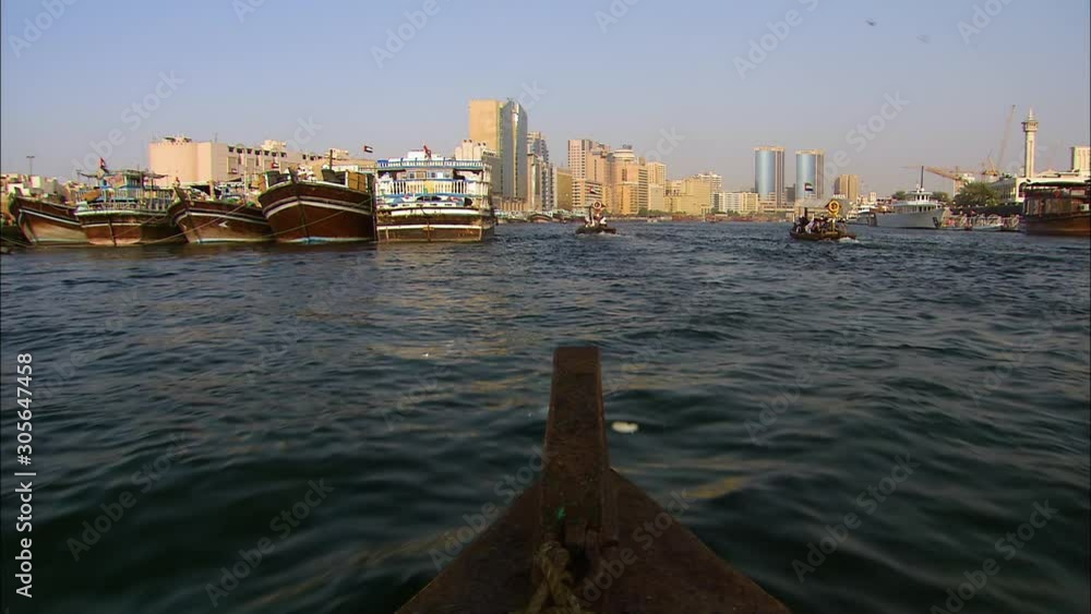 Wide low-angle push-in shot of an Arabian wooden boat sailing on a dark coastal water channel and buildings on both Deira and Dubai Creek sided of the waterway, Dubai Wide low-angle push-in shot of an Arabian wooden boat sailing on a dark coastal water channel and buildings on both Deira and Dubai Creek sided of the waterway, Dubai