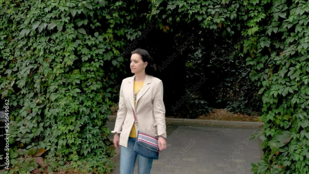 caucasian middle aged woman slim beige jacket jeans walks quickly along a beautiful green alley Cloudy autumn day Camera moves Long shot caucasian middle aged woman slim beige jacket jeans walks quickly along a beautiful green alley Cloudy autumn day Camera moves Long shot