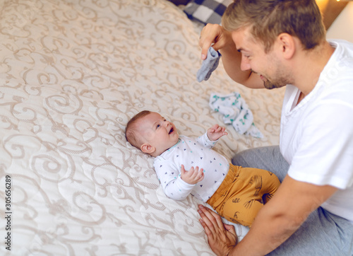 Obraz na płótnie Happy handsome caucasian young dad holding tiny baby socks and preparing to change clothes to his laughing cute 6 months old son
