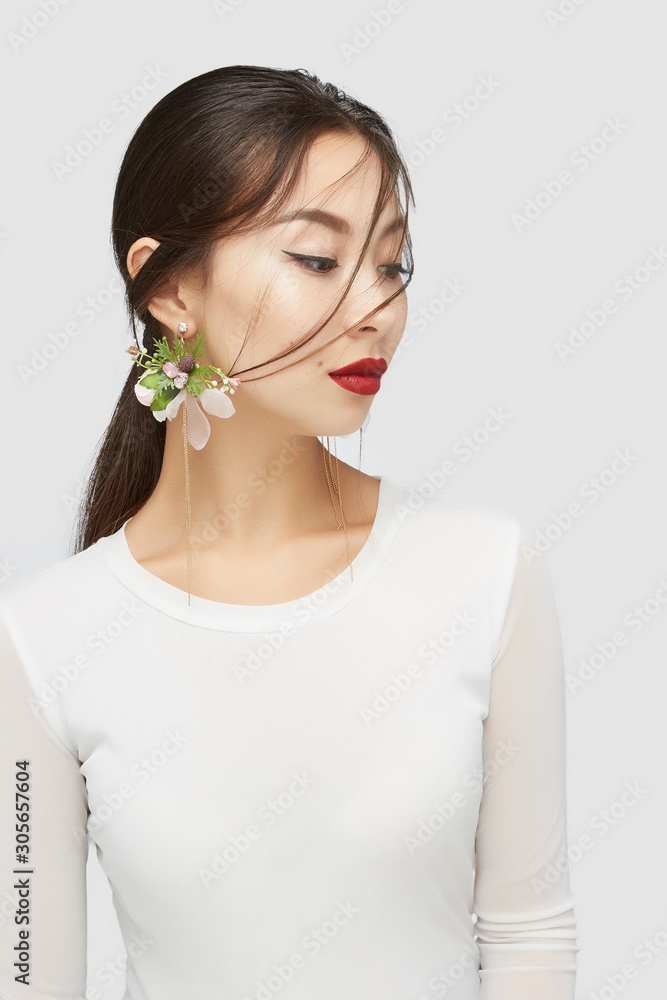 Naklejka premium Medium shot of a young Asian lady in a white blouse and with golden earrings with sparkling crystals and pendants in the form of flower arrangements with gems and golden beads strings.