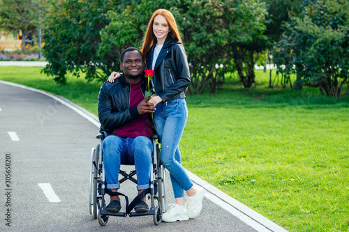 afro american man sitting on wheelchair ,his redhaired ginger girlfriend rolling stroller in autumn park. having romantic date