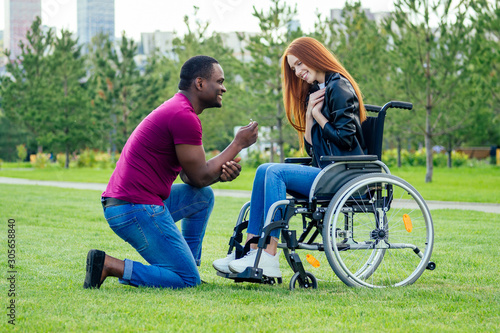 afro american man marriage proposal giving a ring to his redhaired ginger girlfriend.she sitting on wheel chair and surprised and amazed