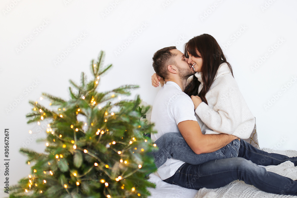 Loving couple. Happy passionate lovers in white sweaters and jeans hug, kiss, roll and fool around on the bed in a white cozy bedroom decorated with a decorative Christmas tree.