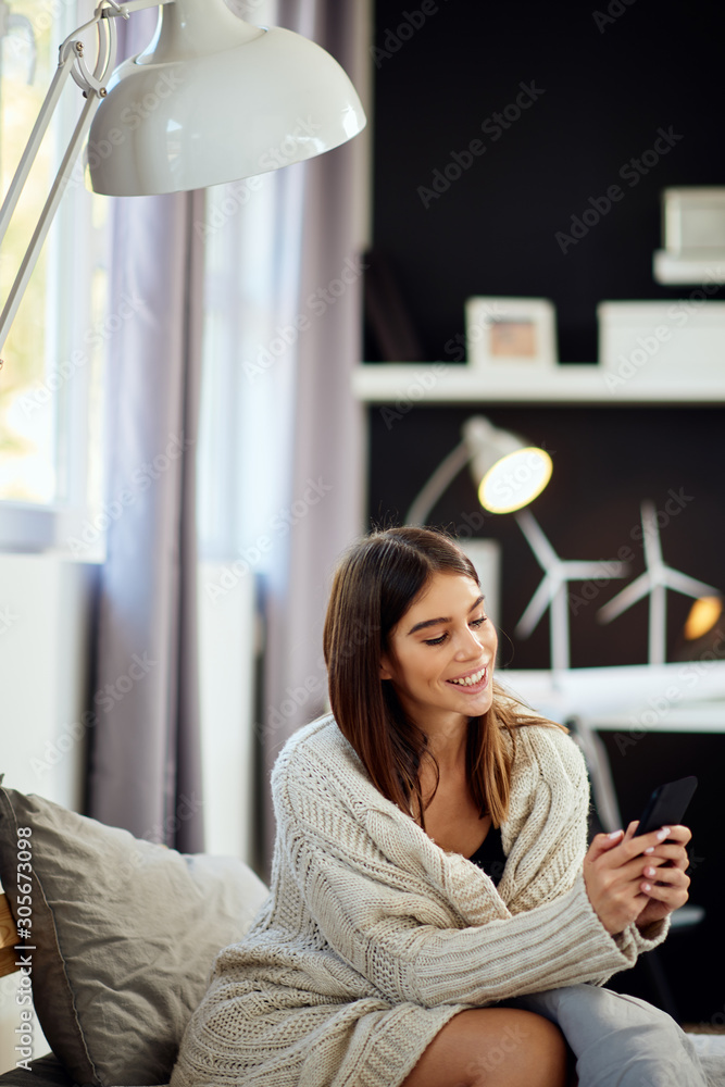 Attractive caucasian brunette dressed in beige sweater sitting on bed in bedroom and using smart phone. Attractive caucasian brunette dressed in beige sweater sitting on bed in bedroom and using smart phone.