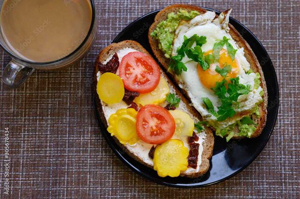 breakfast and brunch. two sandwiches of black bread, with cheese and vegetable filling, lie in a black plate, next to a cup of coffee with milk on a textile background breakfast and brunch. two sandwiches of black bread, with cheese and vegetable filling, lie in a black plate, next to a cup of coffee with milk on a textile background
