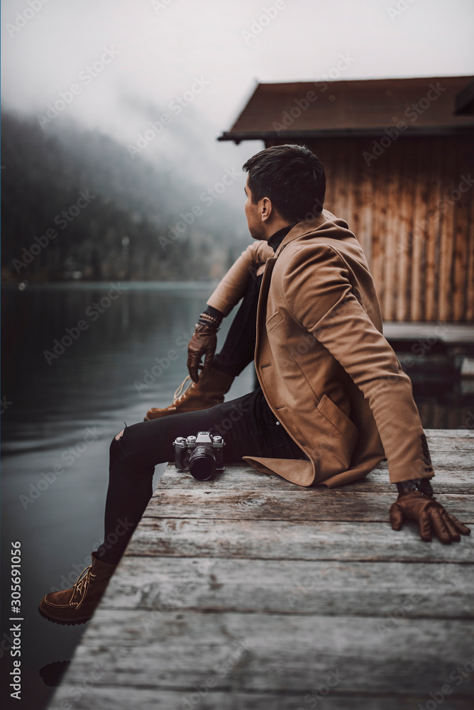 Fototapeta premium Full body shot of young male photographer in brown coat taking pictures with a small mirrorless camera while sitting on a wooden lake house deck in lake Plansee, Austria