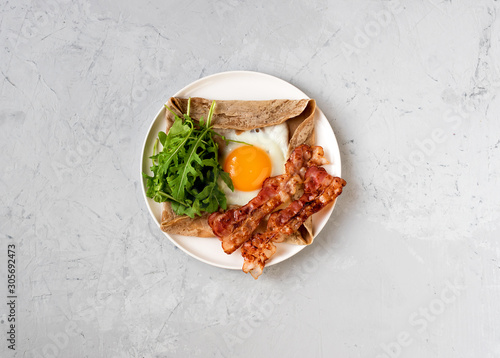 Galette bretonne with fried eggs, arugula and bacon. Plate on a gray background.	