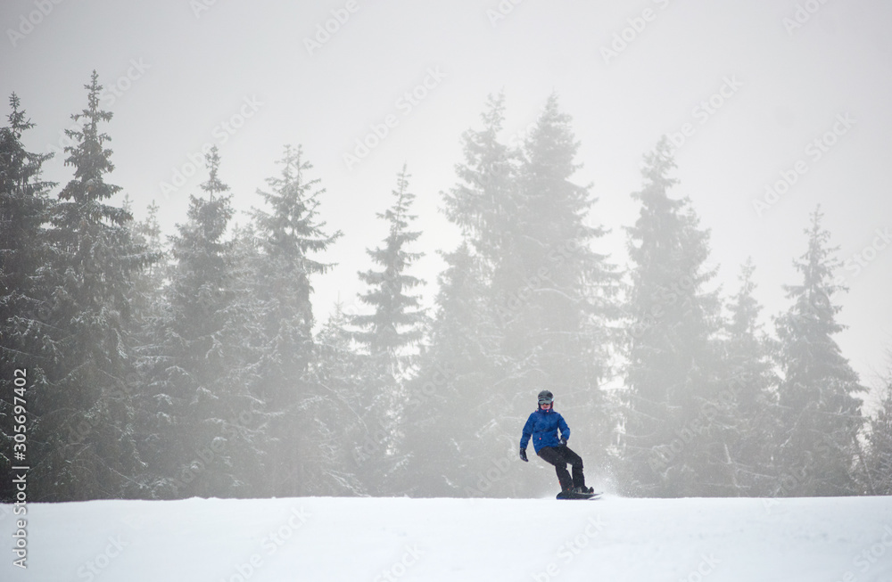 Woman snowboarder sliding on flat snow-covered road. Poor visibility in snowfall. Coniferous forest on background. Copy space. Monochrome view. Freedom, sport, winter activities concept