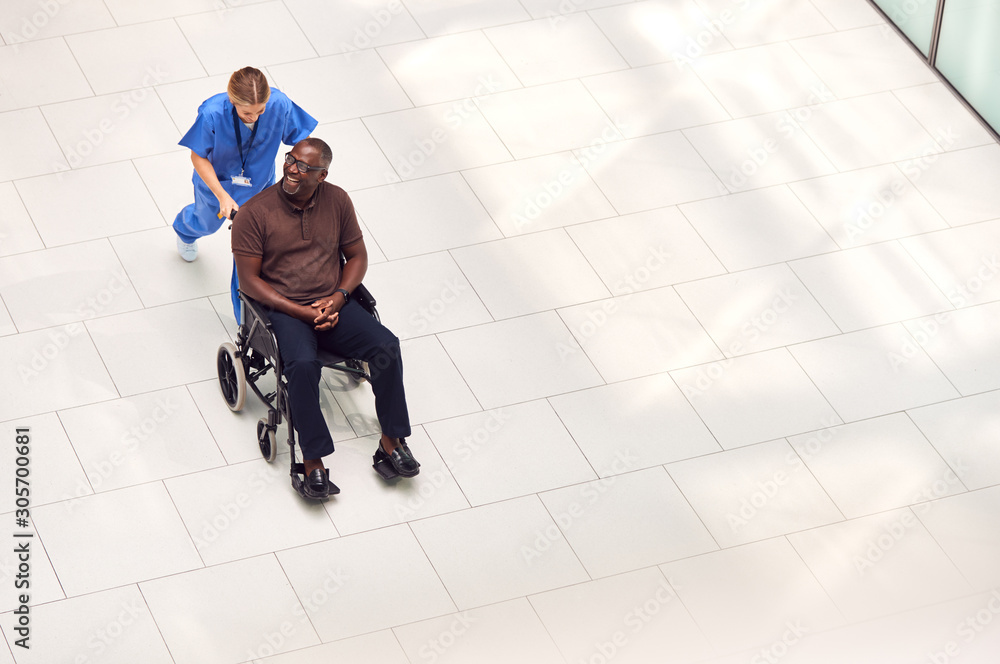 Overhead View Of Nurse Wheeling Male Patient In Wheelchair Through Lobby Of Modern Hospital Building Overhead View Of Nurse Wheeling Male Patient In Wheelchair Through Lobby Of Modern Hospital Building