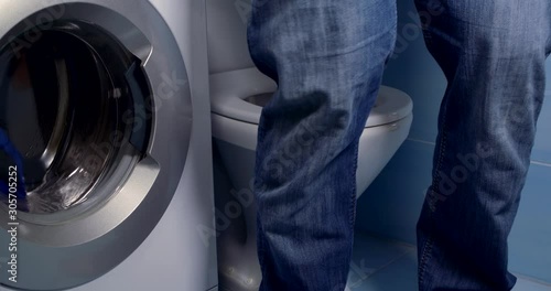 Close-up a man in a shirt lowers his jeans and underpants, sits down on the toilet, rubs his hands, next to it is a washing machine in which the clothes are washed.