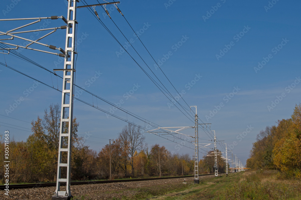 Railway embankment with railroad tracks stretching into distance and perspective above them a power line for the movement of an electric train against a blue sky and trees with green foliage on sides