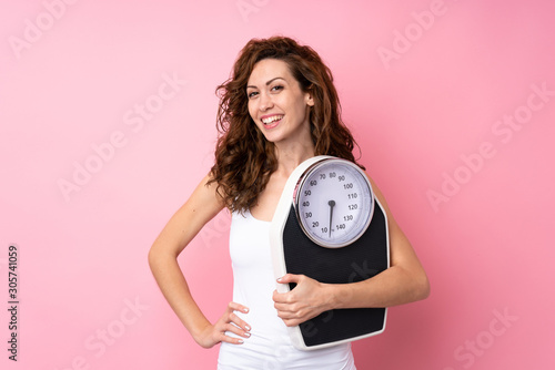 Fotografie Young woman with curly hair holding a weighing machine over isolated pink backgr