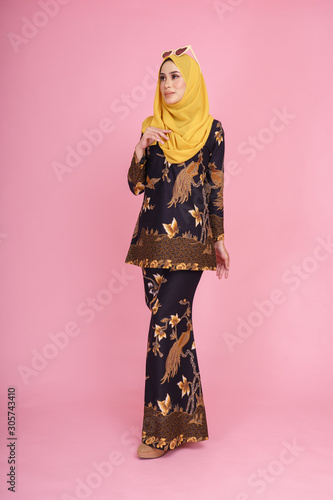 Beautiful female model wearing black "baju kurung" batik design with hijab, a modern lifestyle outfit for Muslim woman isolated over pink background. Eidul fitri fashion and beauty concept.
