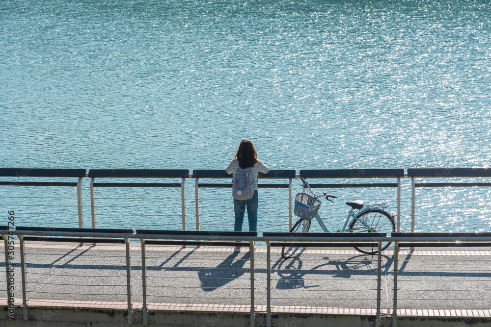 young beautiful girl stand near a bicycle to looking at the view on bike trail at the lake in the morning. Active people. Outdoors