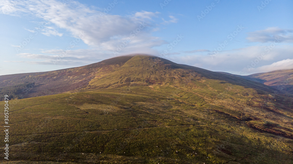 An aerial view of a mountain valley with grassy and stony slopes, trees and mountain range summits under a majestic blue sky and some white clouds An aerial view of a mountain valley with grassy and stony slopes, trees and mountain range summits under a majestic blue sky and some white clouds