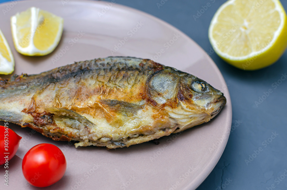 Fried appetizing fish crucian carp with seasoning on a beige plate on a gray background decorated with slices of tomato and lemon. Mediterranean diet. healthy eating concept. Fried appetizing fish crucian carp with seasoning on a beige plate on a gray background decorated with slices of tomato and lemon. Mediterranean diet. healthy eating concept.
