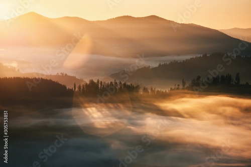 Mountains in fog at beautiful sunrise in autumn in Bucovinas,Romania. Landscape with valley, low clouds, trees on hills, orange sky with clouds at dusk.