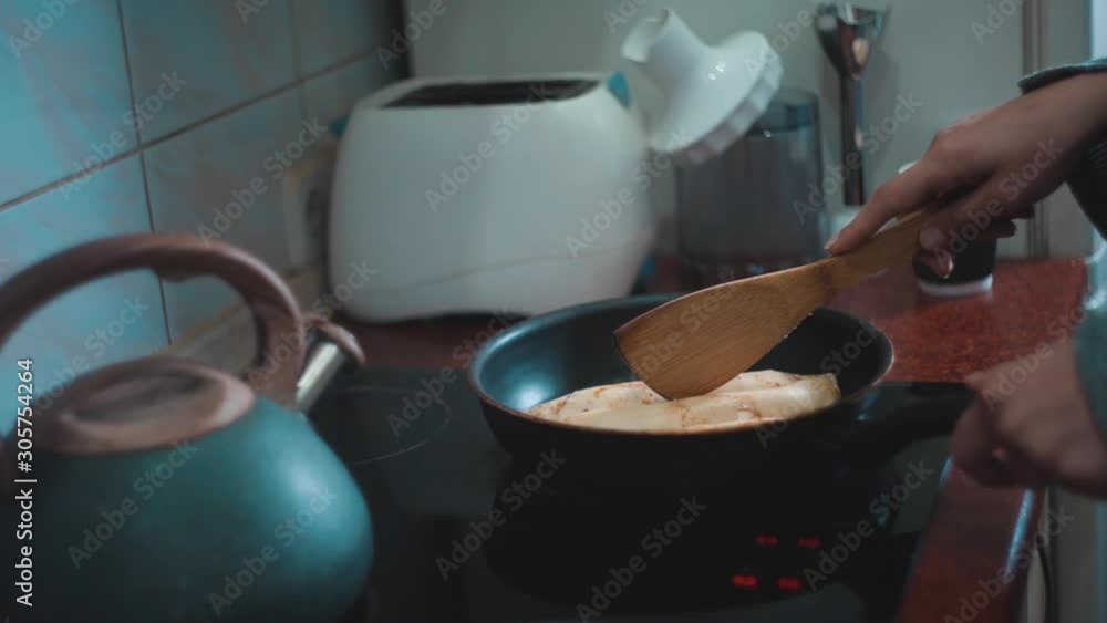 Close up female hands frying rolled pancakes in a pan on an electric stove at home at kitchen. Using a wooden kitchen spatula. Adult girl warming up breakfast Close up female hands frying rolled pancakes in a pan on an electric stove at home at kitchen. Using a wooden kitchen spatula. Adult girl warming up breakfast