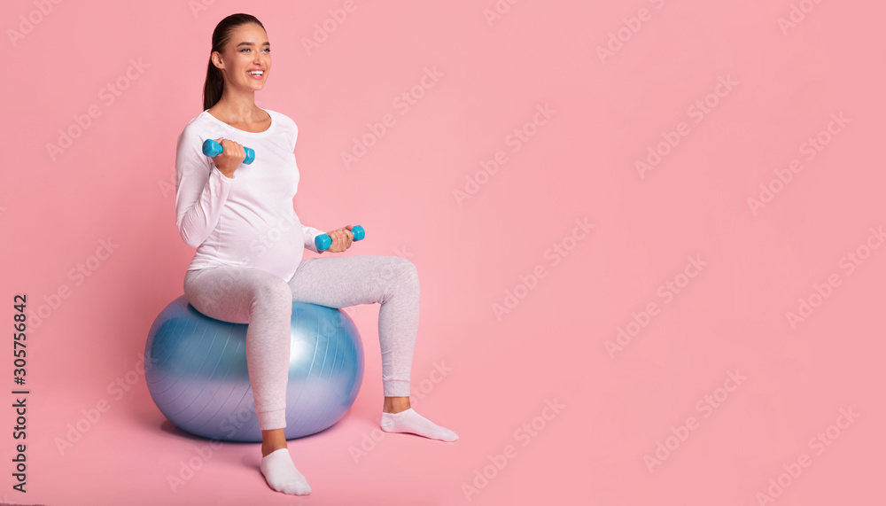 Pregnant Girl Exercising With Dumbbells Sitting On Fitball, Pink Background