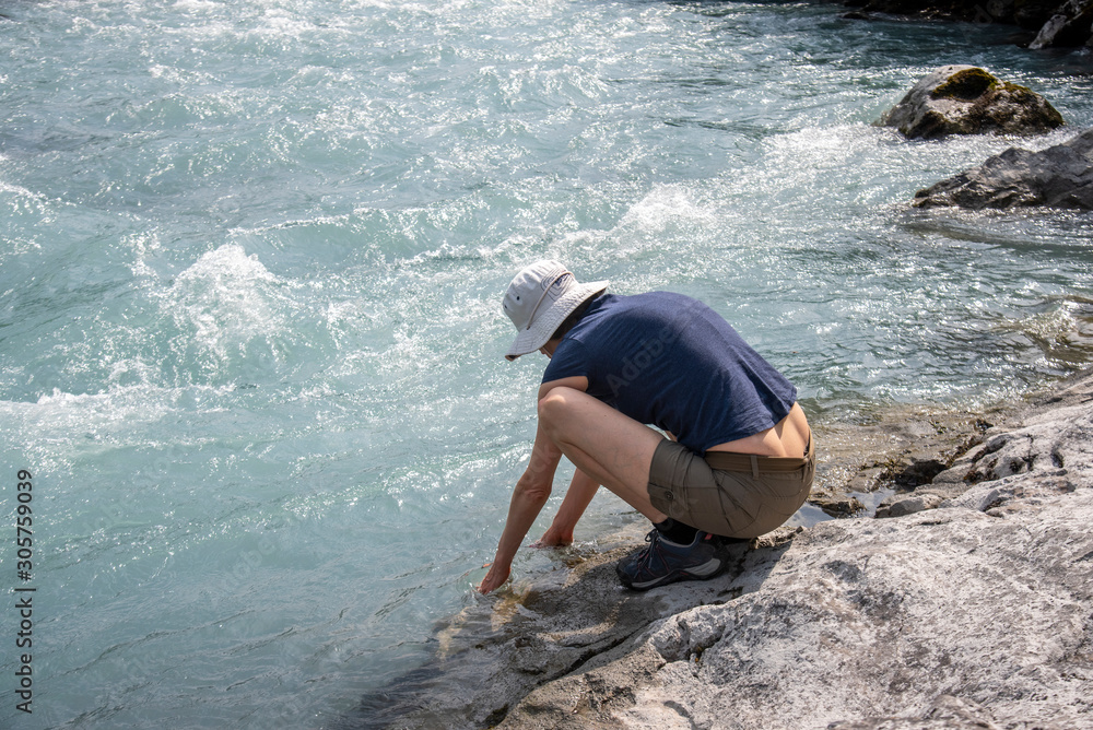 hiking woman on a rock refreshing her hands in the water by the shores of a river with shorts and hat. Sunny summer