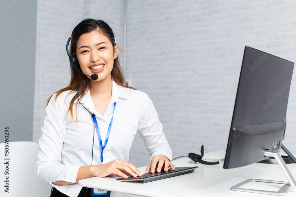 Happy Asian woman with headset smiling and looking at camera while using computer during work in office of call center Happy Asian woman with headset smiling and looking at camera while using computer during work in office of call center