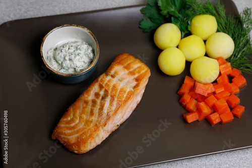 Grilled salmon on a brown rectangular plate with vegetables, potatoes, carrot, lemon, and greens: parsley and dill; with sauce dip. Top view