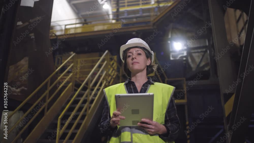 Low angle waist-up arc shot of female factory technician in hard hat and high visibility vest standing in mineral wool insulation manufacturing plant, using tablet, looking around then at camera Low angle waist-up arc shot of female factory technician in hard hat and high visibility vest standing in mineral wool insulation manufacturing plant, using tablet, looking around then at camera